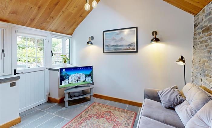 A comfortable living room with a grey sofa, exposed stone wall, flat-screen TV, and a large window and door overlooking a garden. The space features a
